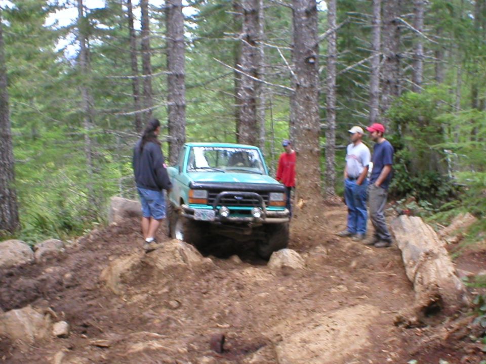 Teal Ford Ranger with brush guard dropping into rutted dirt hole while four people spot