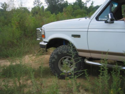 1996 Ford Bronco - A white Ford Bronco with off-road modifications is seen from the front quarter angle on a dirt path surrounded by vegetation.