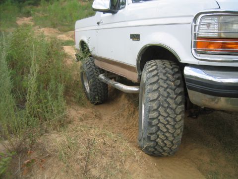 1996 Ford Bronco - A white 1996 Ford Bronco with off-road tires navigates a dirt trail with overgrown vegetation.