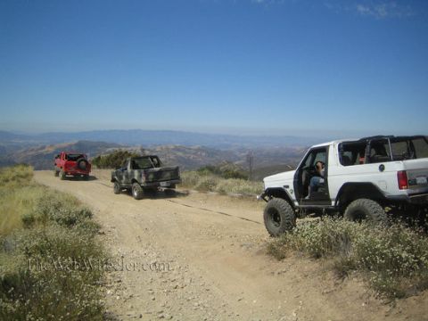 1985 Ford Bronco - A white 1985 Ford Bronco is being towed by a camouflage truck up a dusty mountain road, with other vehicles visible in the distance.