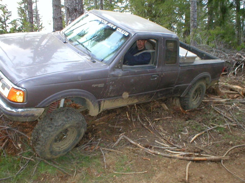 Gray Ford Ranger extended-cab 4x4 with red coil springs flexing over brush in the woods