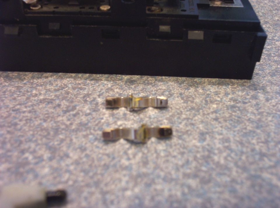 Two metal clips on a speckled surface, with a black electronic component behind them. "SUPERMOTORS www.supermotors.net" text is visible