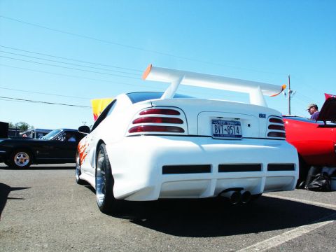 2001 Mercury Grand Marquis - A modified white car with a prominent spoiler is parked next to other vehicles under a clear blue sky.