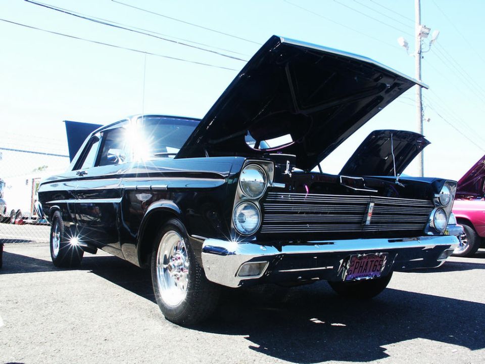 Black 1964 Mercury Comet with hood open, showing chrome grille and dual headlights. Another car is visible to the right