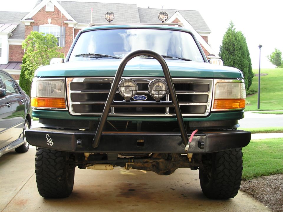Front view of a green Ford Bronco with black stinger bumper, mesh-covered driving lights and mud-terrain tires