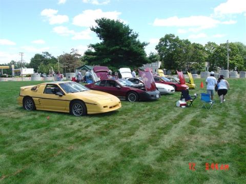 1984 Pontiac Fiero - Several Pontiac Fieros are parked on a grassy field under a partly cloudy sky.