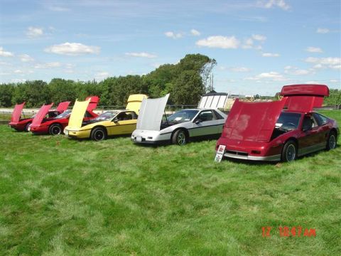 1984 Pontiac Fiero - Several Pontiac Fieros are lined up on a grassy field with their hoods open under a partly cloudy sky.