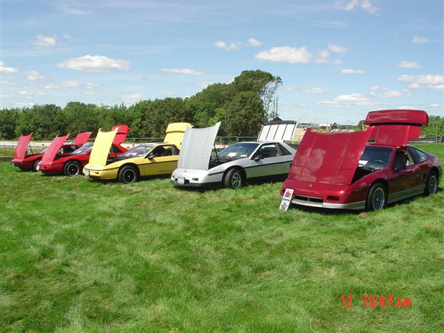 Row of red, yellow, and silver Pontiac Fieros with hoods open on a grassy field