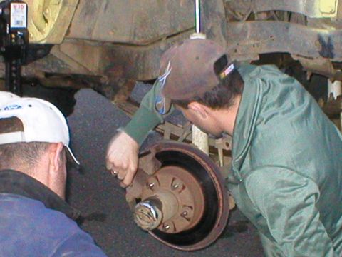 1978 Ford Bronco - Two individuals examine the brake system of an unidentified vehicle, focusing on the rotor and caliper.
