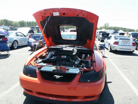 2001 Mercury Grand Marquis - A car show features an orange Ford Mustang with its hood up, revealing the engine bay. Several other vehicles are parked nearby.