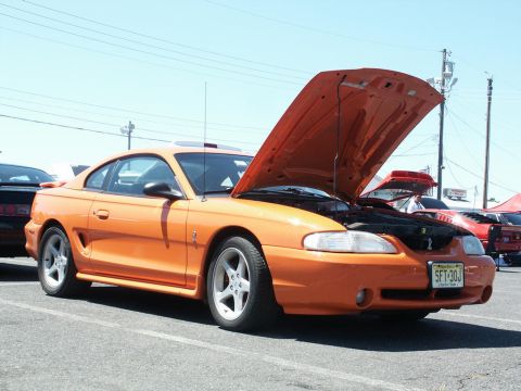 2001 Mercury Grand Marquis - An orange Ford Mustang is parked with its hood up, showing the engine bay. Other cars are visible in the background.