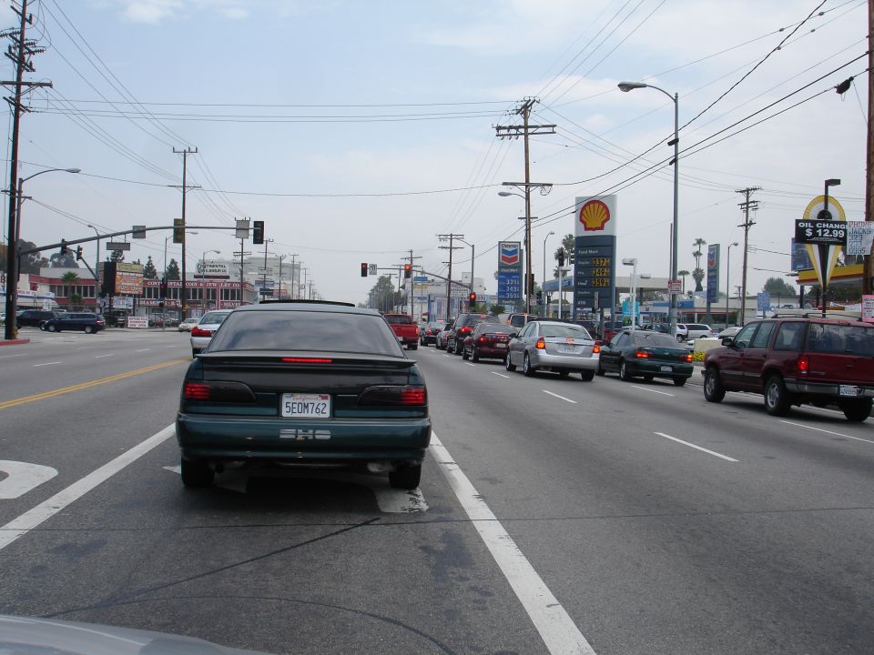 Green sedan with a spoiler and a license plate reading 5EOM762, stuck in traffic on a street with gas stations and power lines