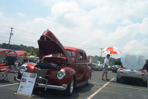1985 Ford Crown Victoria LTD - A classic car show features a red coupe with its hood open, alongside other vehicles and attendees.