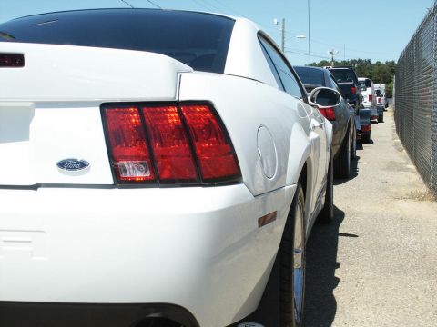 2001 Mercury Grand Marquis - A white Ford Mustang is parked in a line of vehicles next to a chain-link fence.