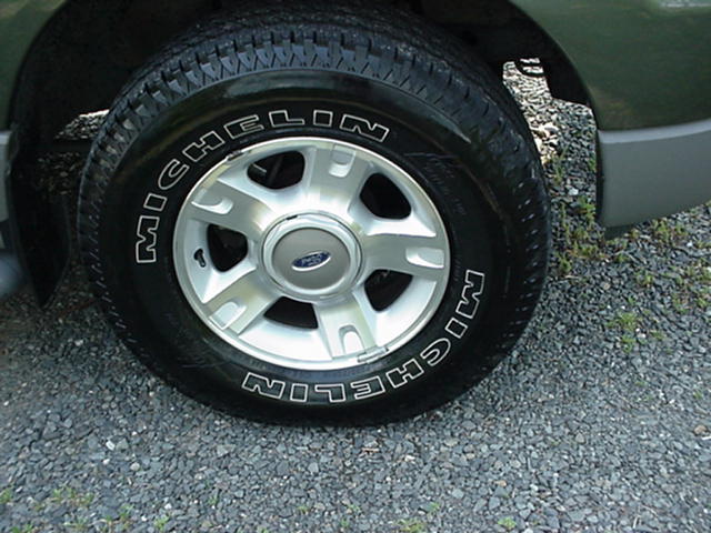 Michelin white-letter tire on silver alloy wheel with Ford center cap, green vehicle on gravel