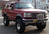 1994 Ford Bronco - Small low-resolution thumbnail of a red lifted Ford Bronco shot from the front three-quarter driver side on a residential street, with a pe…