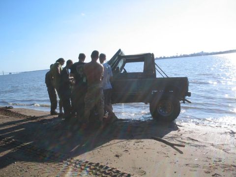 1990 Ford Bronco - A group of people gather around a dark SUV partially submerged in the water at the edge of a sandy beach.