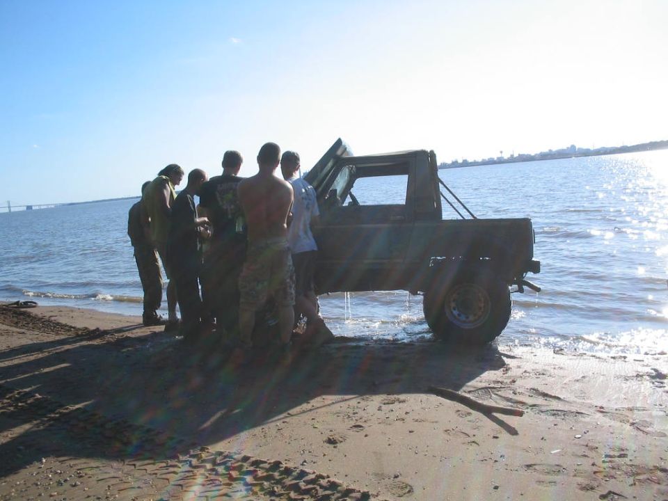 Side view of a dark SUV partially in water, surrounded by people on a beach