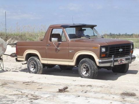 1985 Ford Bronco - This brown Ford Bronco, seen from a 3/4 front angle, is parked on a sandy area with dunes in the background.