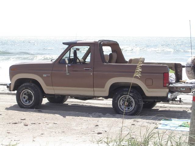 Brown 1985 Ford Bronco parked on a sandy beach with the ocean in the background