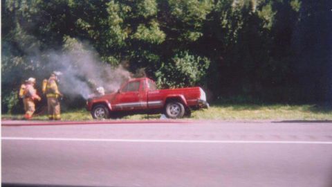 1993 Ford Taurus - Firefighters attend to a red pickup truck that is on fire by the side of the road.