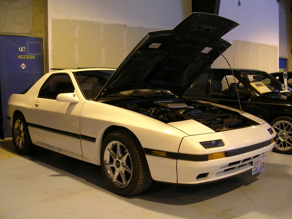 White Mazda RX-7 with hood open, chrome wheels, and a license plate reading 557-UDM