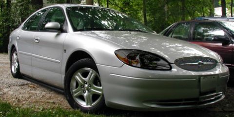 1993 Ford Taurus - A silver sedan is parked on a gravelly surface with grass and trees in the background. Another vehicle is visible behind it.
