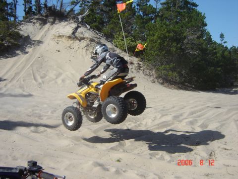 1993 Ford Bronco - A rider on a yellow ATV soars through the air over a sandy hill, with trees and flags in the background.