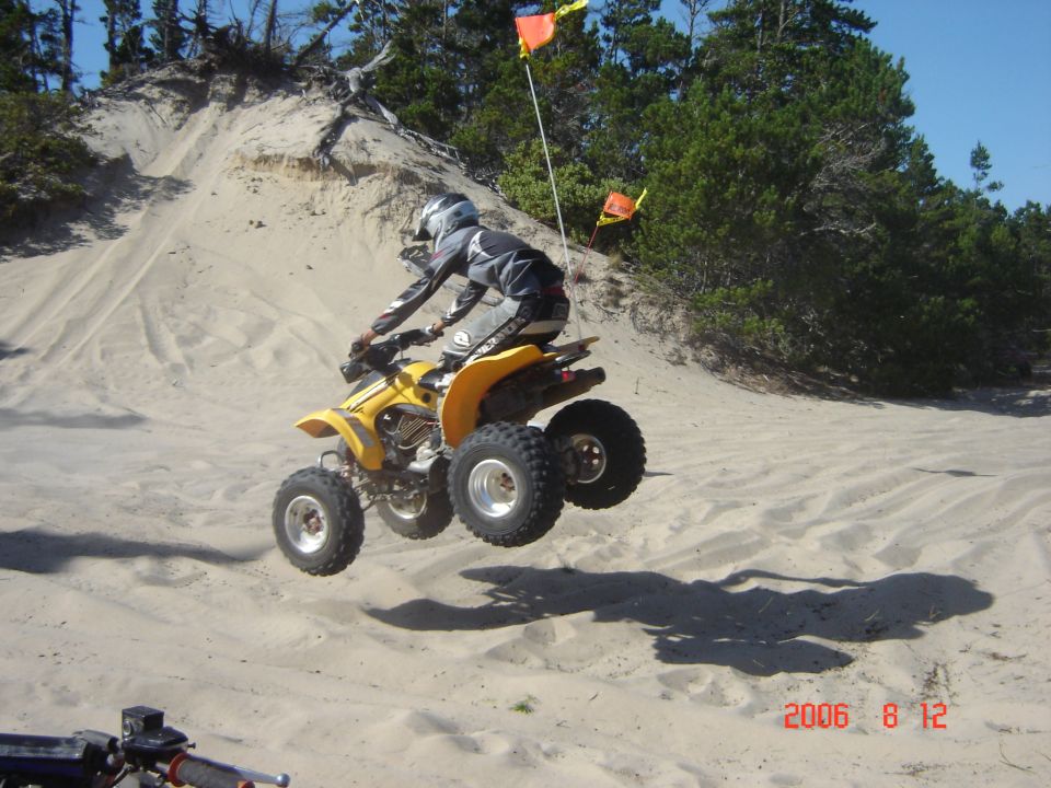 Yellow ATV mid-air over a sand dune, rider wearing helmet and gear. Another ATV is partially visible in the foreground