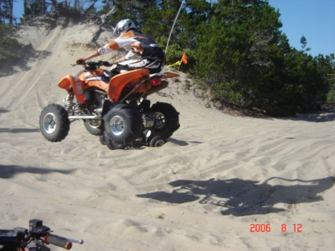 1993 Ford Bronco - A quad bike rider takes flight over a sandy dune, with trees and a clear blue sky overhead.