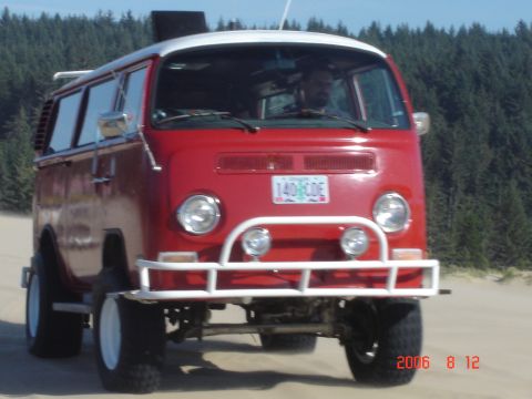 1993 Ford Bronco - A red Volkswagen Type 2 van, modified for off-road use with large tires, drives on a sandy terrain near a forest.