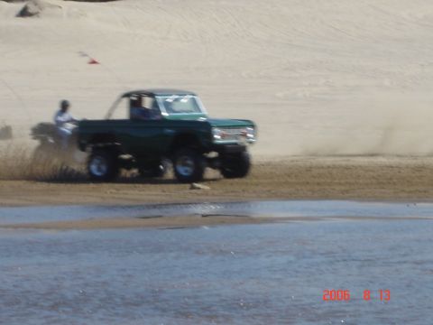 1993 Ford Bronco - A lifted Ford Bronco kicks up sand as it drives along the shore, with a small body of water in the foreground.
