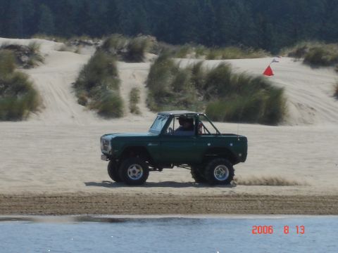 1993 Ford Bronco - A green Ford Bronco drives on a sandy beach with dunes and vegetation in the background.