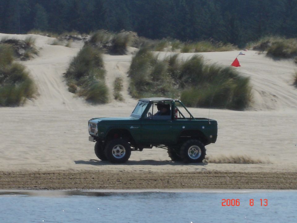 Green Ford Bronco with roll cage driving on sand, water in foreground