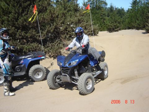1993 Ford Bronco - Two ATVs are on a sandy dune with trees in the background. One rider is on the ATV, the other stands next to theirs.