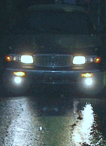 1985 Mercury Grand Marquis - The front of a car is illuminated by its headlights and fog lights at night, with reflections on a wet road surface.