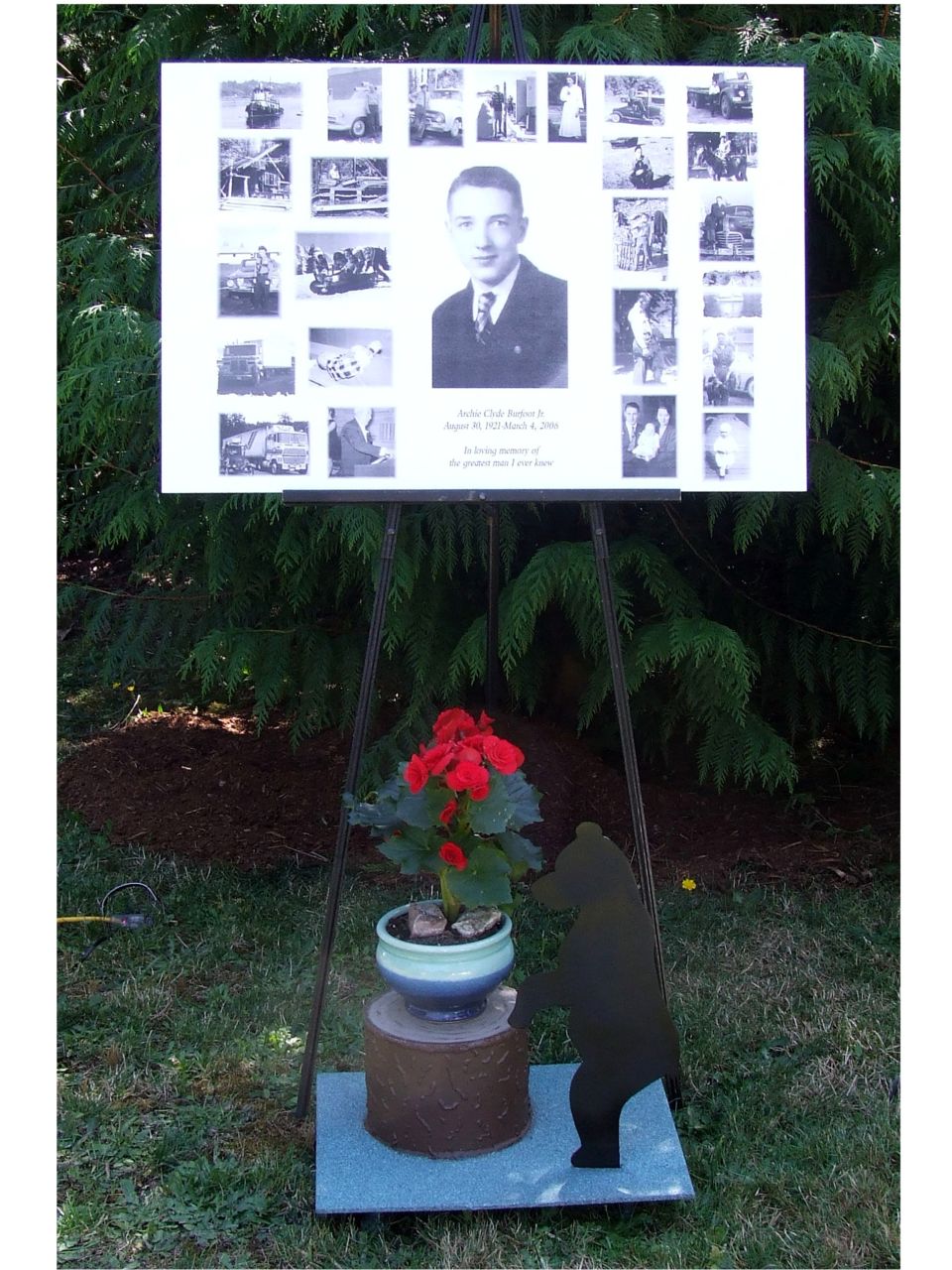 Memorial display board with a portrait and many small photos, set up outdoors with a potted plant and a bear silhouette