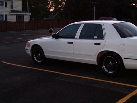 1999 Ford Crown Victoria - Side view of a white sedan parked in a lot during sunset. It has black wheels and a spotlight.