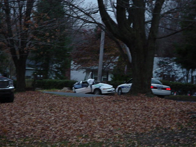 Damaged white sedan with doors open, parked next to a police car on a leaf-covered road