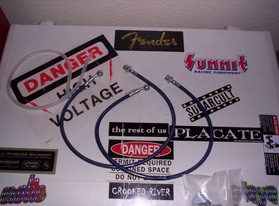Two blue brake lines with metal fittings lie on a white surface covered in stickers, including 'Danger High Voltage', 'Fender', and 'Summit Racing Equipment'
