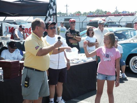 2001 Mercury Grand Marquis - A man speaks into a microphone at an outdoor event, with people and vehicles gathered around.