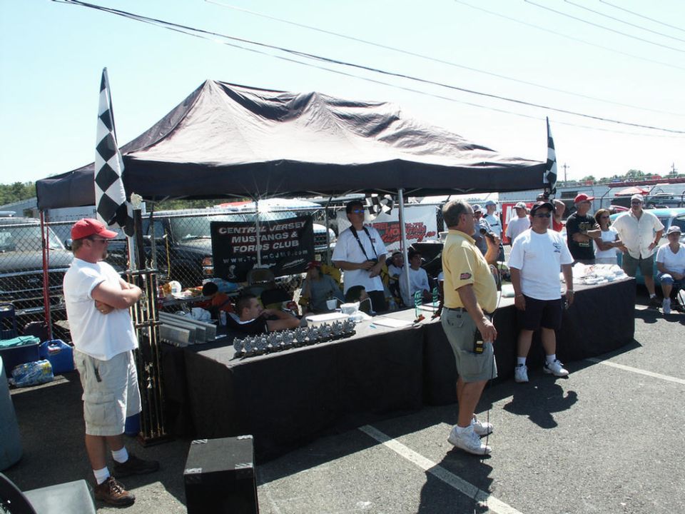 people gathered under a tent at an outdoor event, with cars visible in the background
