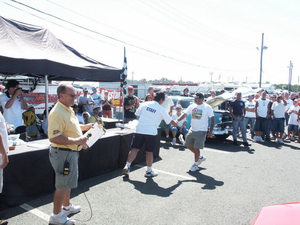 People gathered at an outdoor car show, with a tent and awards table in the foreground