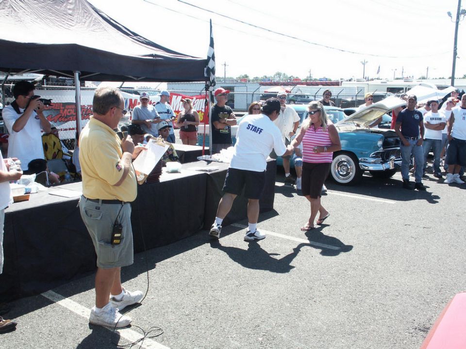 People gathered around a table at an outdoor event, with a classic car in the background. A person in a staff shirt is handing something to a woman