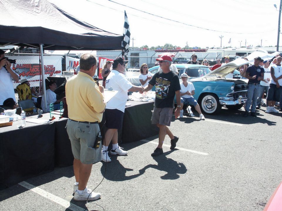 People at a car show, one man shakes hands with another near a table with a checkered flag. A blue classic car is parked behind them with its hood up