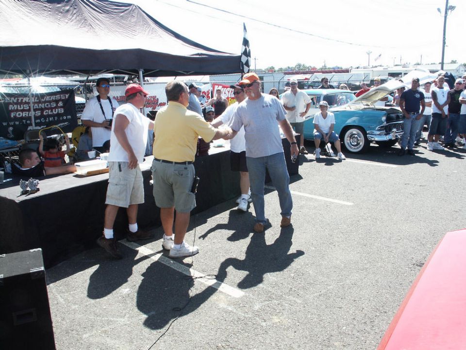 People shaking hands at a car show, with a turquoise 1955 Ford Fairlane in the background