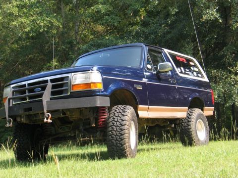 1995 Ford Bronco - A lifted blue Ford Bronco with large tires sits on a grassy incline, surrounded by trees.