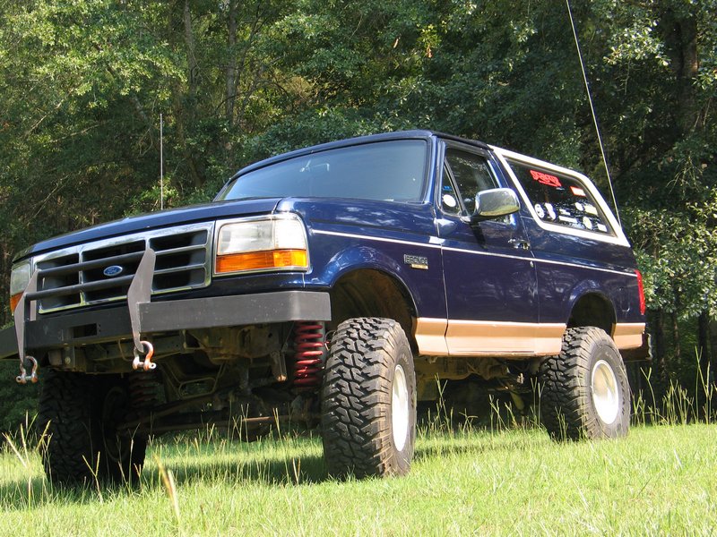 Lifted blue and tan Ford Bronco on mud tires parked in a grassy field, 3/4 front view