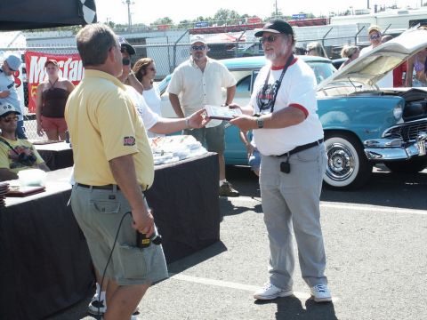 2001 Mercury Grand Marquis - Two men exchange an award at an outdoor event, with a vintage car on display.
