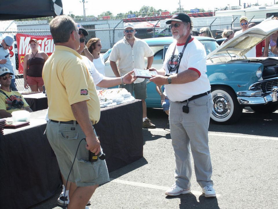 Man in yellow shirt hands award to man in white shirt, blue classic car with hood up in background